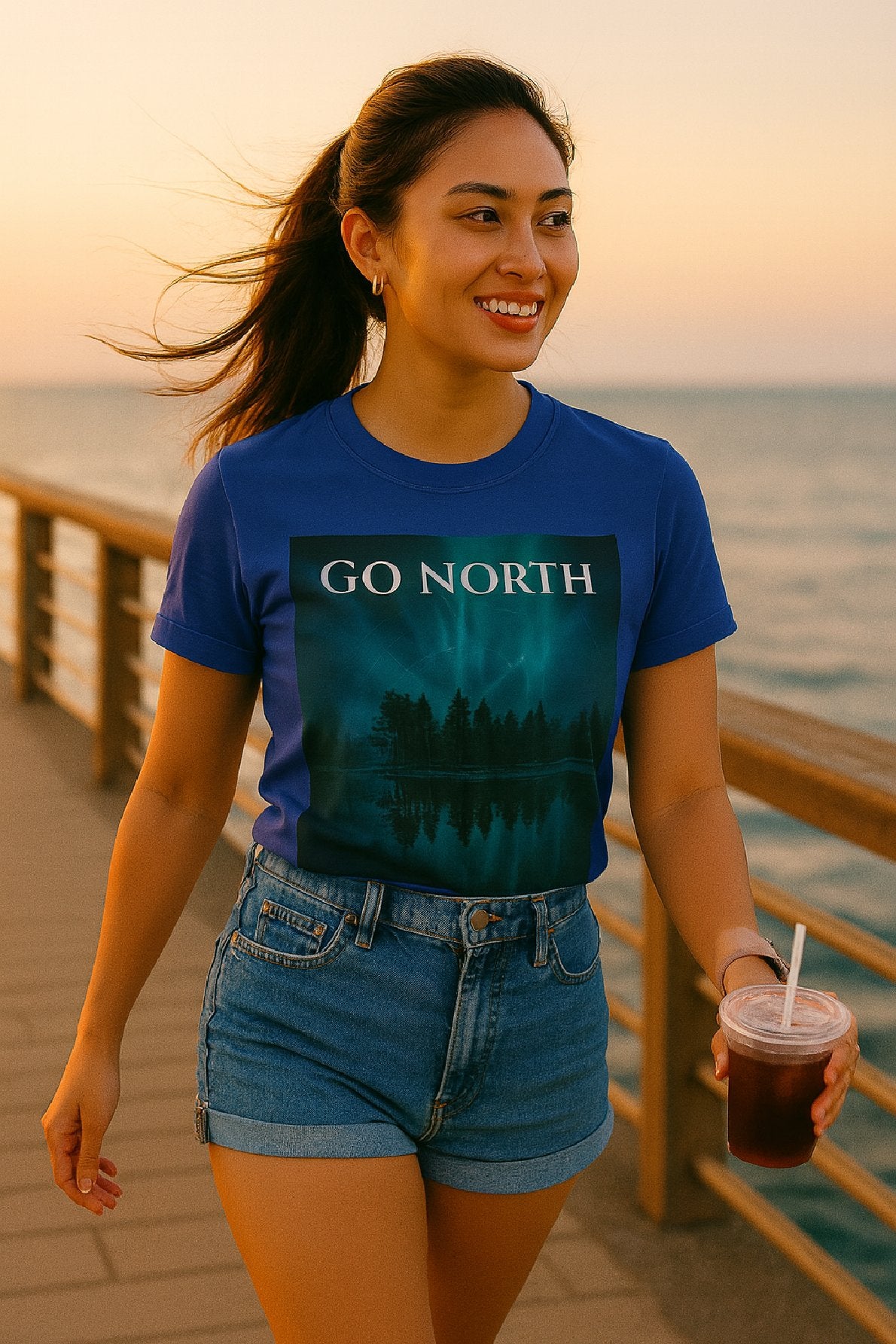 Woman wearing a blue t-shirt with 'GO NORTH' text by the ocean at sunset