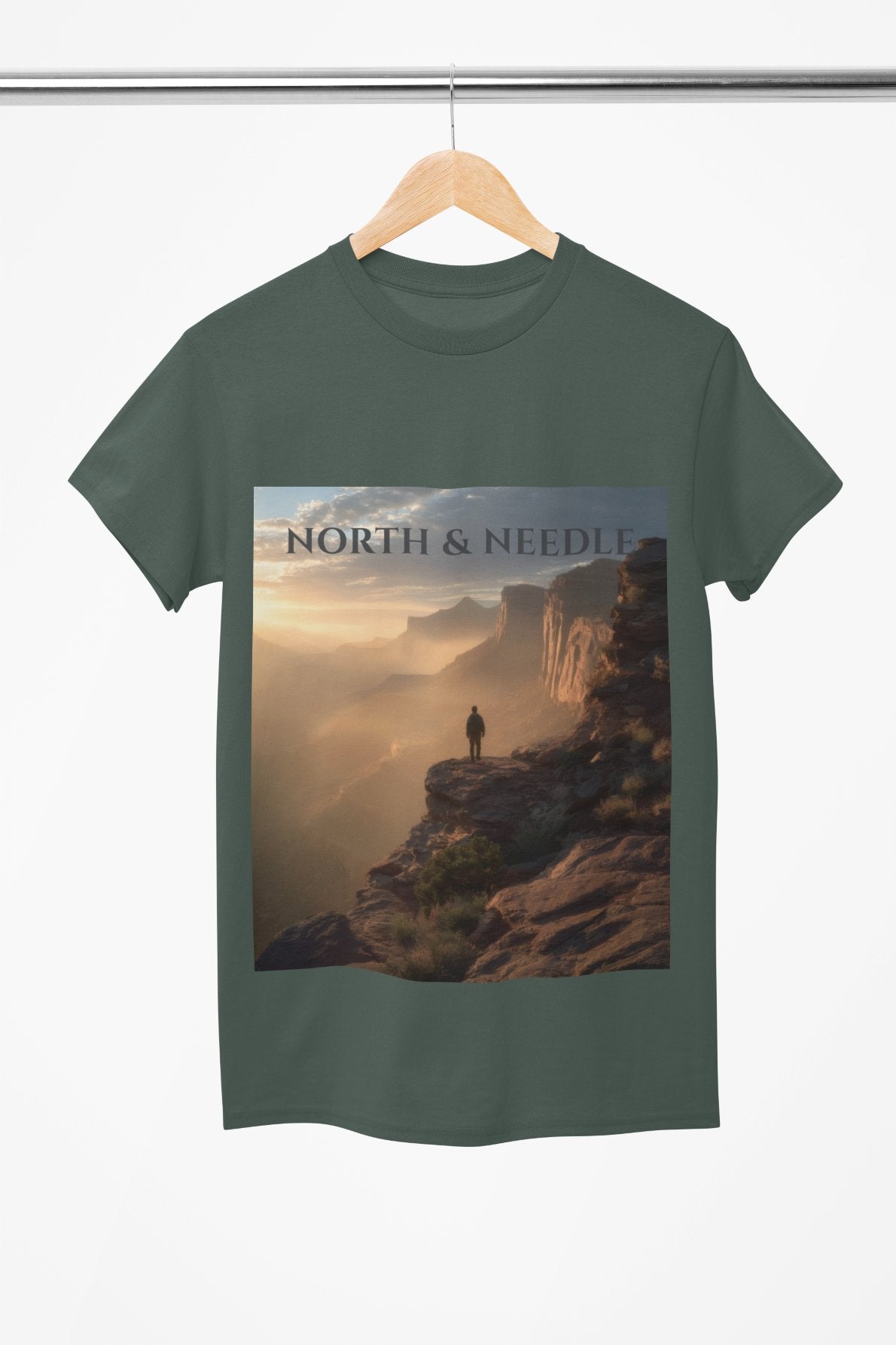 Green t-shirt with a scenic design of a person on a cliff and 'North & Needle' text, hanging on a wooden hanger.
