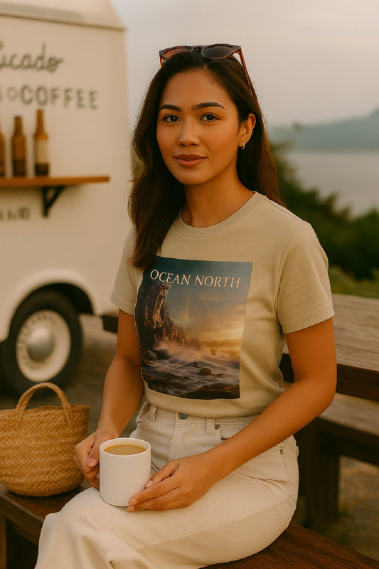 Woman sitting outdoors holding a coffee cup, wearing an 'Ocean North' t-shirt with a scenic design.
